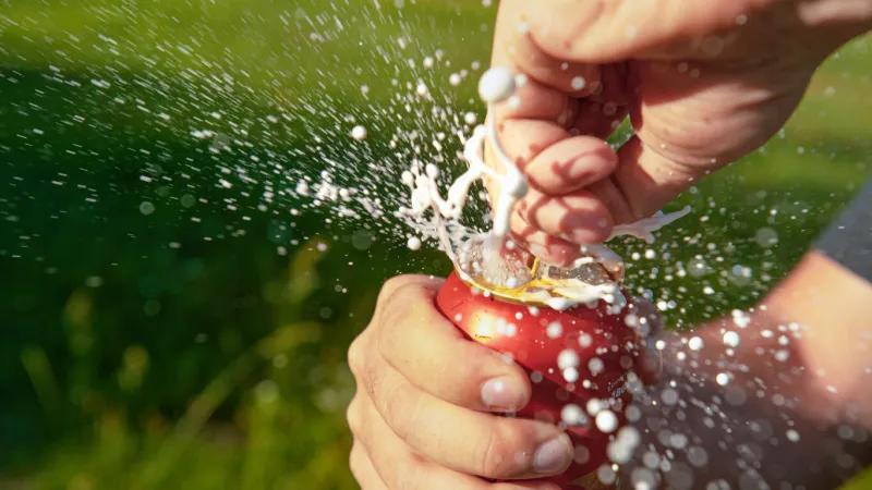 close up, dof  unrecognizable thirsty man opens a can of shaken up beer during a fun summer picnic unfortunate guy spills refreshing cold beer while pulling the tab of a large red aluminium can