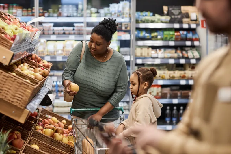 side view portrait of black young woman with little girl shopping for groceries in supermarket and buying fruits