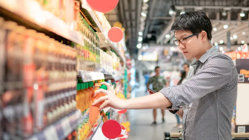 asian man choosing orange juice in supermarket using smartphone to check shopping list male shopper with shopping cart selecting beverage bottle product in grocery store