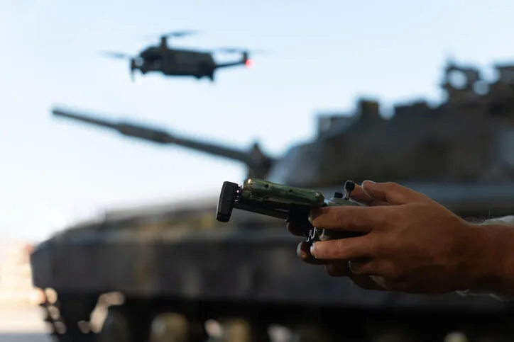 a man with a uav control panel in his hands, a drone flying above the tank in the background in nature help of reconnaissance drones in modern warfare