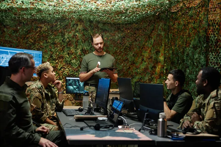 young confident officer of surveillance room using tablet during report and looking at one of colleagues while standing in front of them