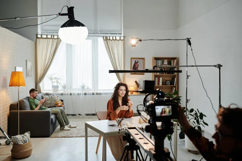 happy young female blogger sitting by table in loft apartment during video record of commercial being shot by cameraman