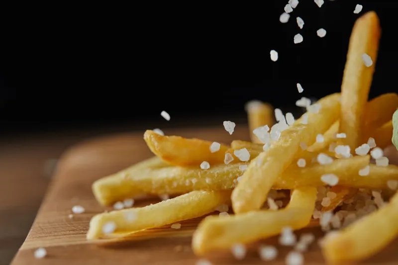close up of salt falling on fresh golden french fries on wooden chopping board