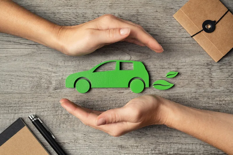 close up of hands of young woman protecting icon of green eco-friendly car over wooden table top view of woman hands protecting electric car with care, mobility car insurance and green automotive business concept