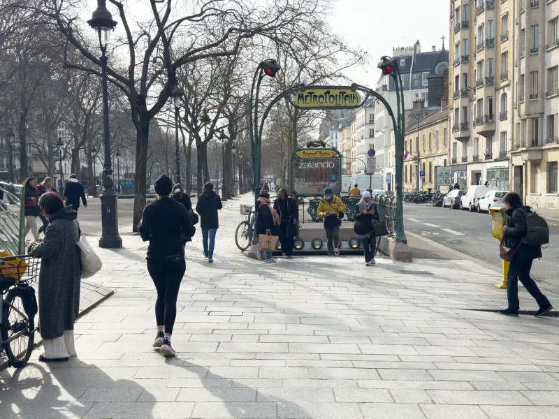 paris,france-15022025 city dwellers walk by a vintage metro entrance, surrounded by trees and shops, enjoying the crisp air of a sunny afternoon in a lively urban setting