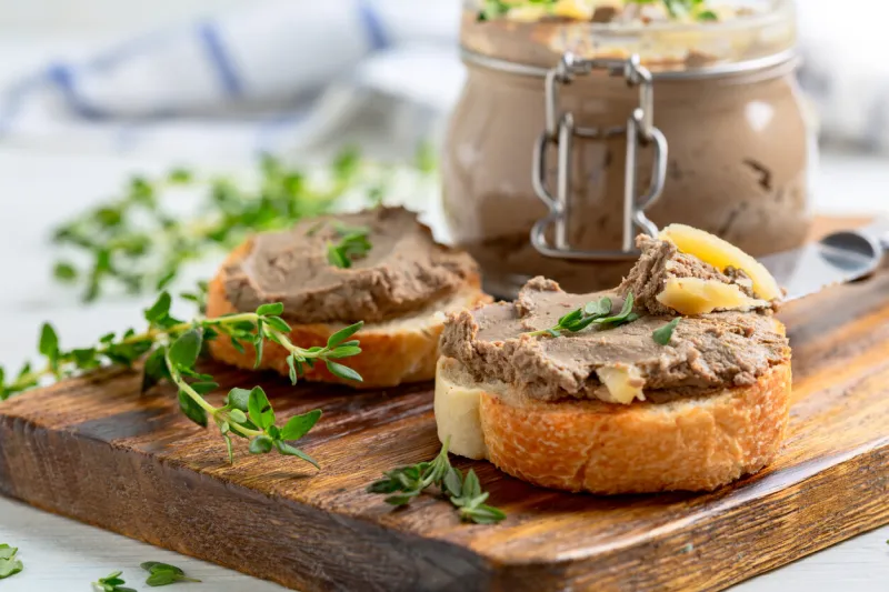 slices of toasted baguette with turkey pate with thyme and a jar of pate are served on a wooden service board concept of delicious homemade food