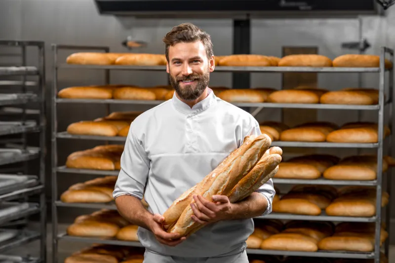 handsome baker in uniform holding baguettes with bread shelves on the background at the manufacturing