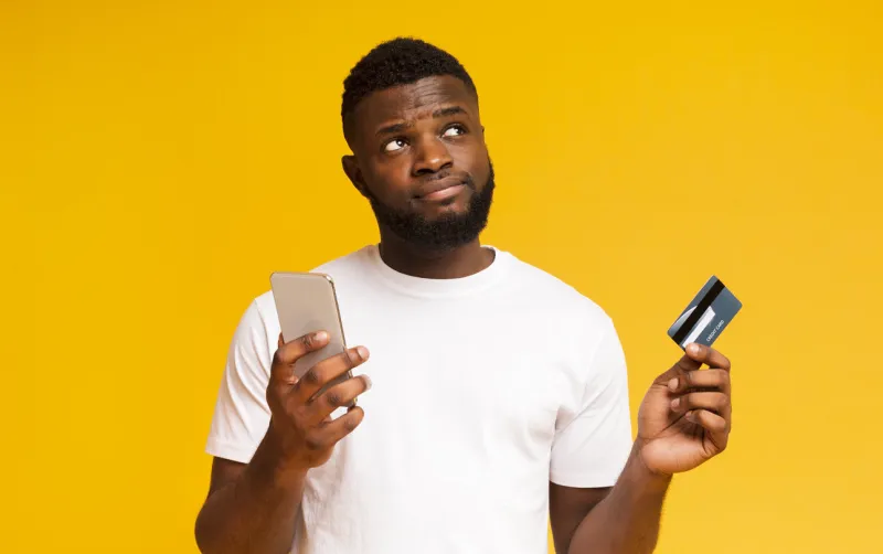 do i longer need a credit card hesitant african american man holding smartphone and card, looking upwards