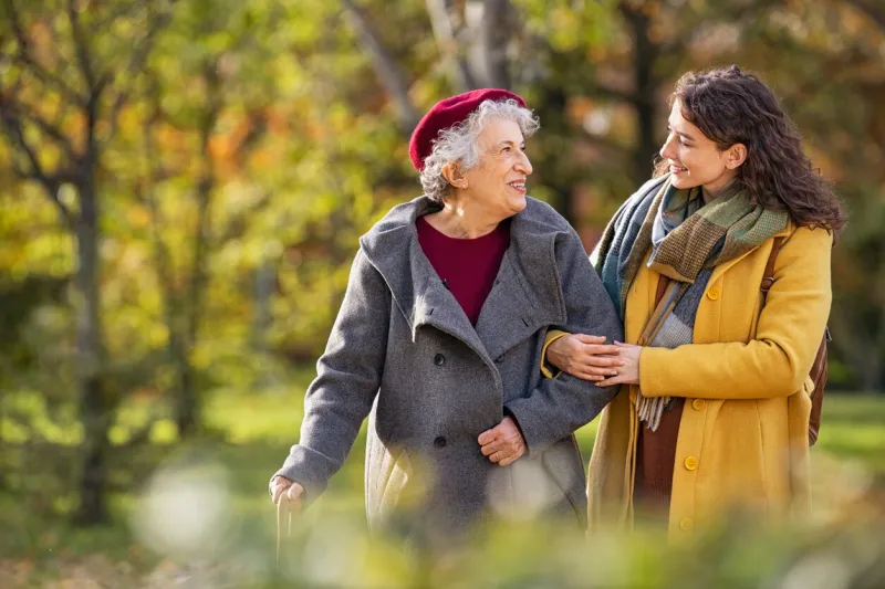 young woman in park wearing winter clothing walking with old grandmother happy grandma wearing coat walking with lovely girl outdoor with copy space smiling lovely caregiver and senior lady walking in park during autumn and looking at each other
