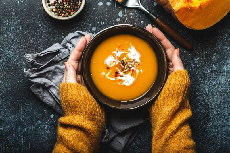 female hands in yellow knitted sweater holding a bowl with pumpkin cream soup on dark stone background with spoon decorated with cut fresh pumpkin, top view autumn cozy dinner concept