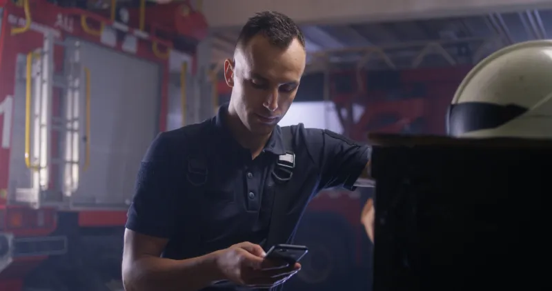 male firefighter texting message on smartphone and speaking with coworker during break on fire station