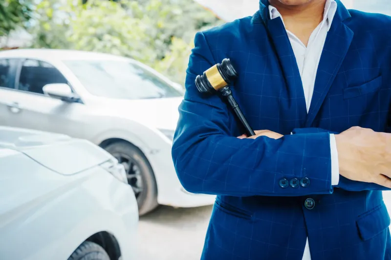 businessman in a suit and a lawyer, holding a wooden gavel, stand in front of a car, discussing legal aspects like citations, liability, and negligence related to a traffic violation