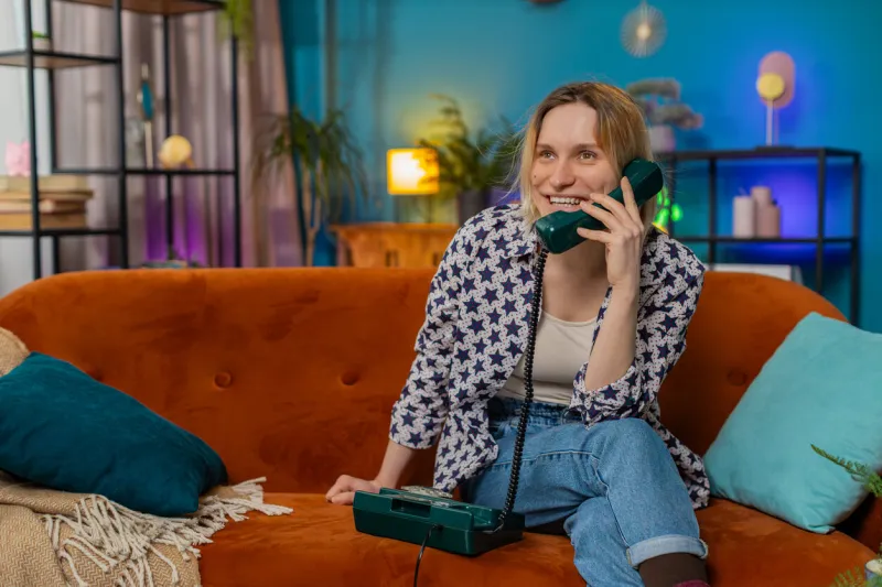 smiling young woman making wired telephone call conversation with friends sitting on couch at home in room happy excited pretty girl enjoying old-fashioned retro phone from 90s talking in living room