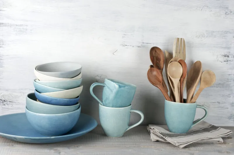 simple rustic handmade blue crockery against white wooden wall  dish, stack of bowls mugs and wooden cooking utensils set