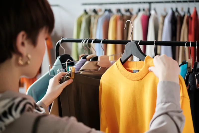 rear view of young woman looking at clothes on rack in her hands and choosing a new style for herself in the clothes store