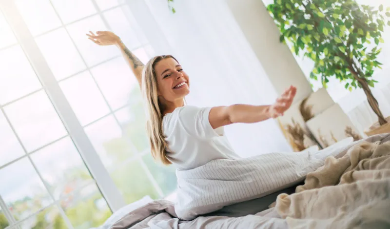 cute smiling woman stretches joyfully on a bed, bathed in natural light from large windows the bedroom is bright and cozy, with plants and soft bedding, evoking a sense of comfort and happiness