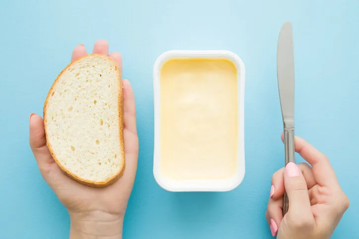 woman's hands holding slice of white bread and knife opened plastic pack of light yellow margarine on pastel blue desk preparing breakfast point of view shot closeup top view