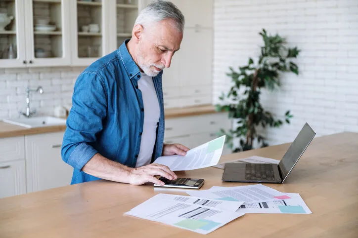 focused middle aged man making calculations of utility payment holding paper bill invoice in hand at table serious businessman doing paperwork for paying taxes online using laptop at home