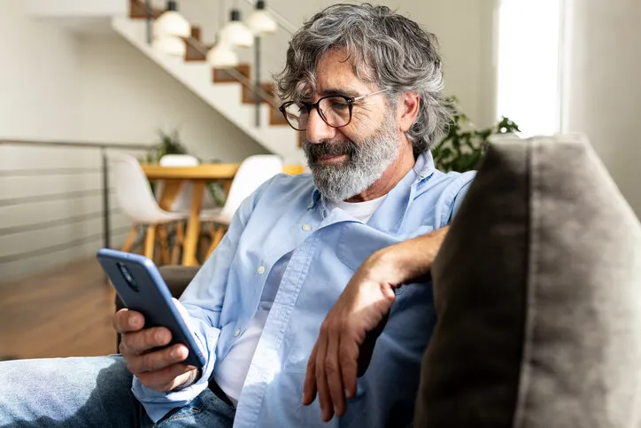 mature man relaxing at home, sitting on the sofa sending text message with mobile phone lifestyle concept