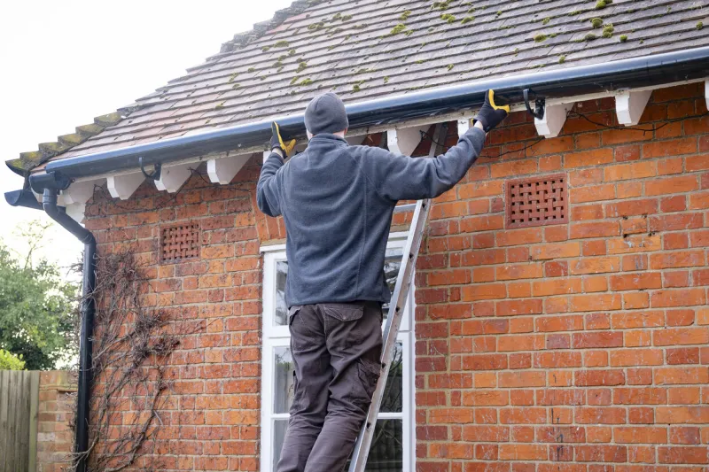 man on a ladder fixing fitting a rain gutter on the eaves of a uk house