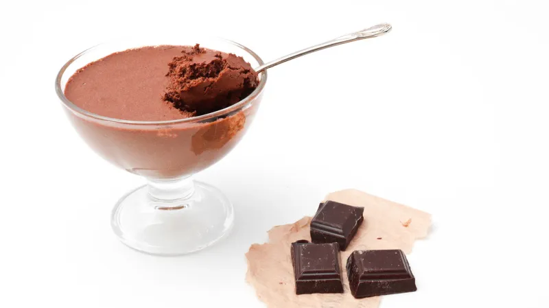 transparent bowl with chocolate mousse and teaspoon and three pieces of dark chocolate isolated on a white background