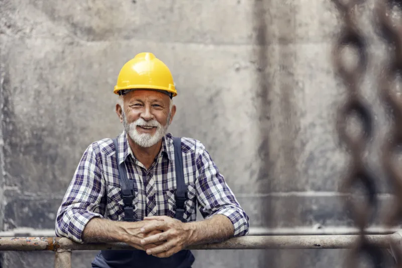 a smiling, senior worker with a helmet on his head, leans on the railing in the factory and takes a break from work in heavy industry a senior worker in a factory