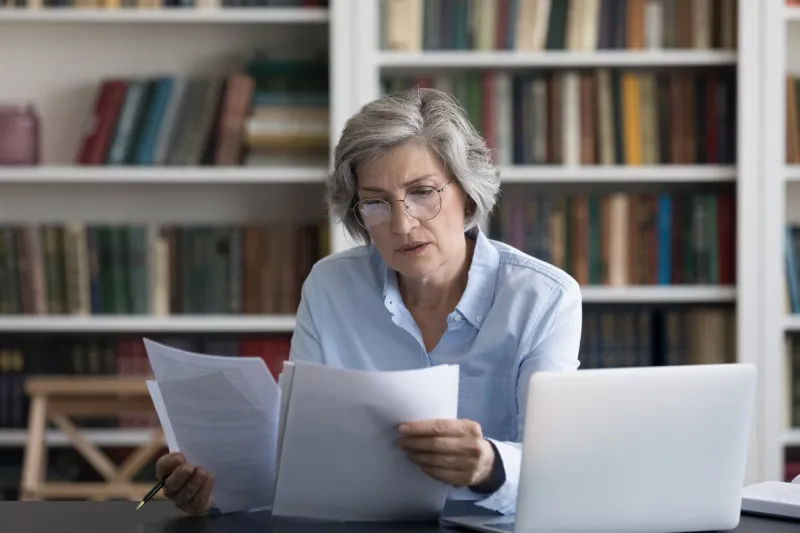 serious thoughtful middle-aged woman in glasses looks worried read news in formal document sit at workplace desk with wireless computer older female review paper letter, learns report feels concerned