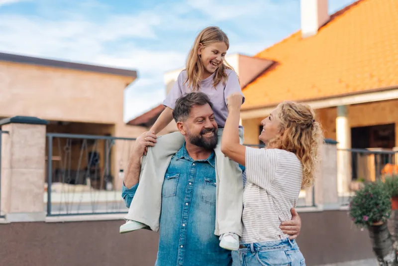 smiling family with daughter on father's shoulders having fun in front of their house