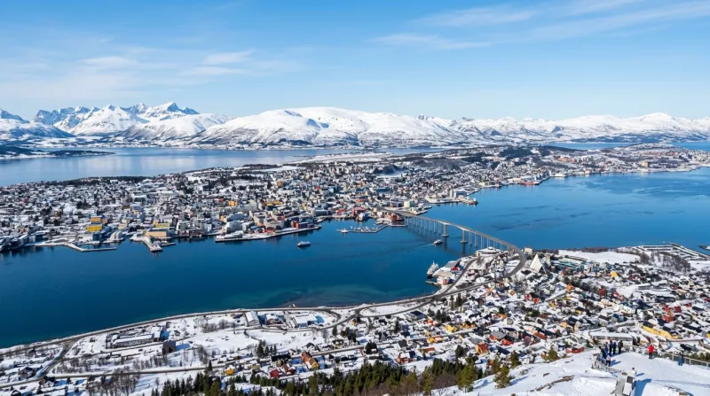 Une photo de la ville de Tromsø en Norvège entourée de fjords et de sommets enneigés sous un ciel se