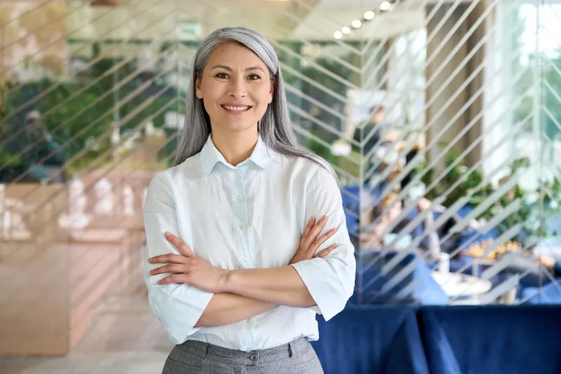 happy smiling confident middle aged asian older senior female leader businesswoman standing in modern office workplace looking at camera arms crossed business successful executive concept portrait