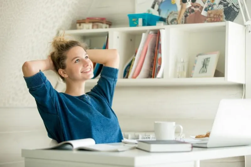portrait of an attractive woman at the table with cup and laptop, book, notebook on it, arms crossed back to her head, relaxed pose bookshelf at the background concept photo