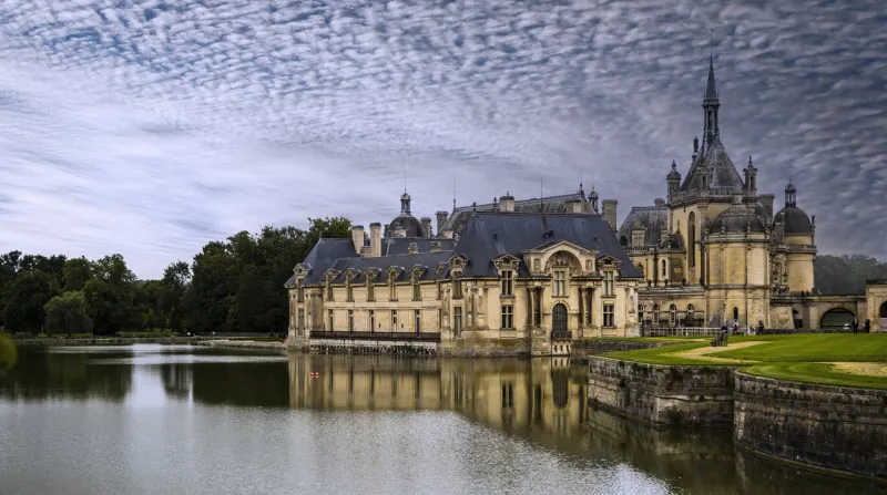 panoramic view of the château de chantilly reflected in the water of the river with light clouds in the sky