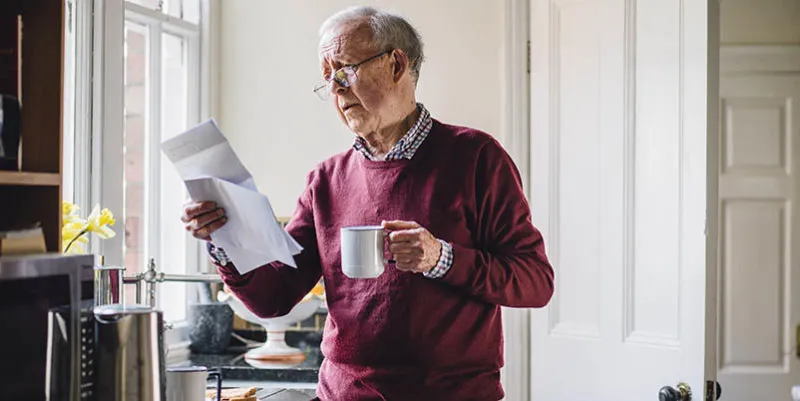 senior man is standing in the kitchen of his home with bills in one hand and a cup of tea in the other he has a worried expression on his face