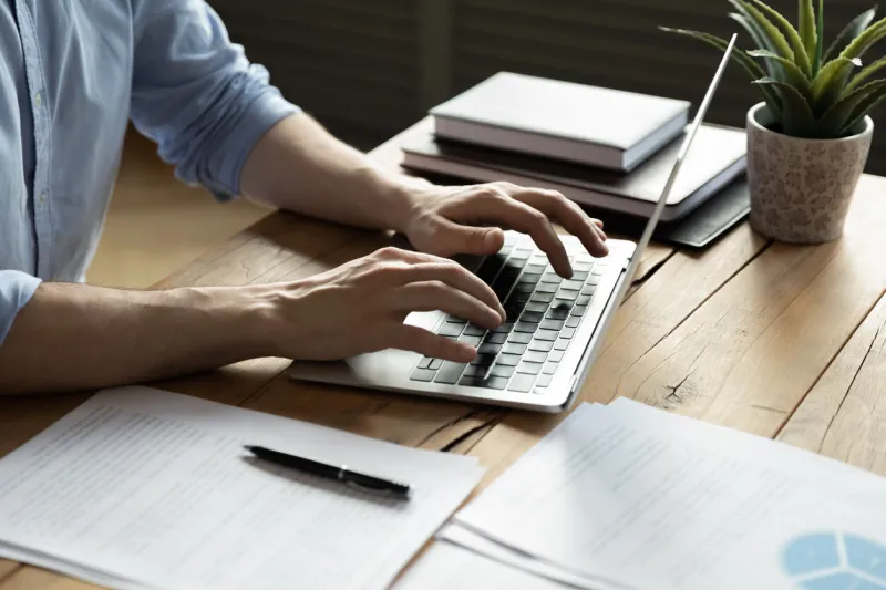 close up businessman using laptop, typing on keyboard, sitting at wooden desk with documents, writing email, accountant writing financial report, busy student studying online, searching information
