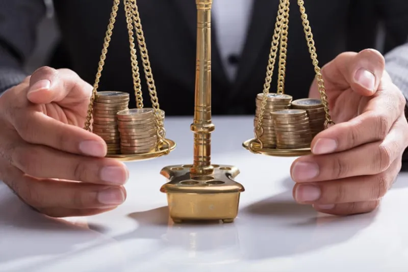 close-up of a businessperson's hand covering stacked coins on golden weighing scale