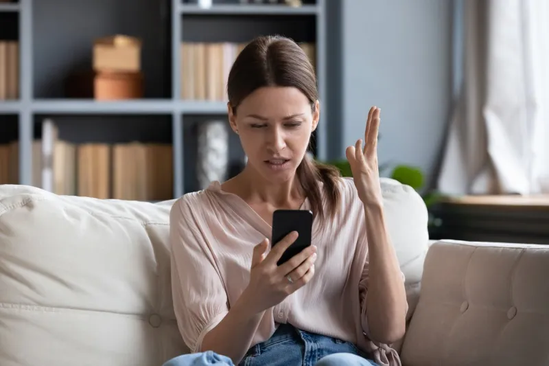 confused angry woman having problem with phone, sitting on couch at home, unhappy young female looking at screen, dissatisfied by discharged or broken smartphone, reading bad news in message