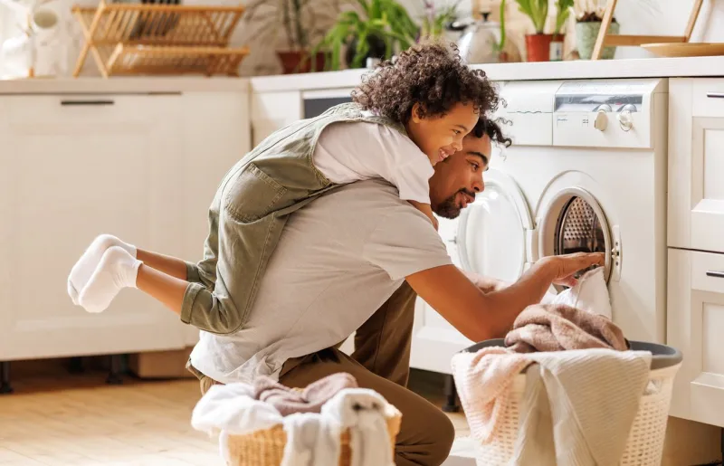 side view of black child in casual clothes with curly hair smiling and embracing dad loading washing machine during household routine in morning at home
