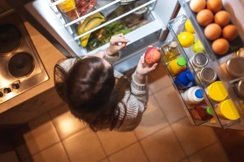 top view of a beautiful young woman standing next to an opened refrigerator door in the kitchen at night, smiling and holding an apple