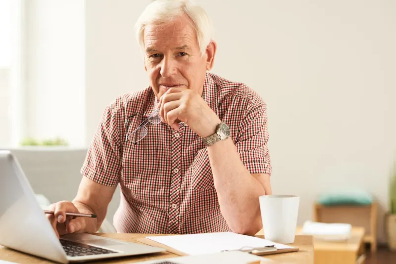 portrait of modern senior man using laptop at home working and looking at camera, smiling
