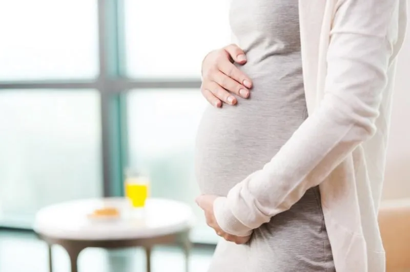 close-up side view image of young pregnant woman touching her abdomen while standing in front of the window and table with orange juice and cookies on it