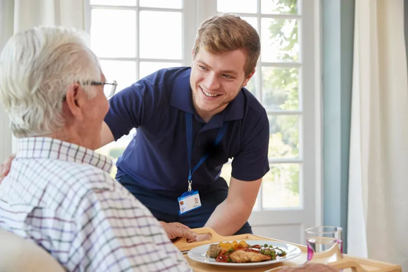 male care worker serving dinner to a senior man at his home