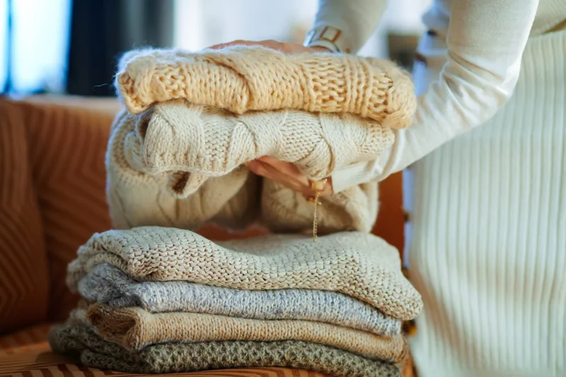 closeup on stylish woman in white sweater and skirt in the modern living room in sunny winter day near couch folding sweaters for storage