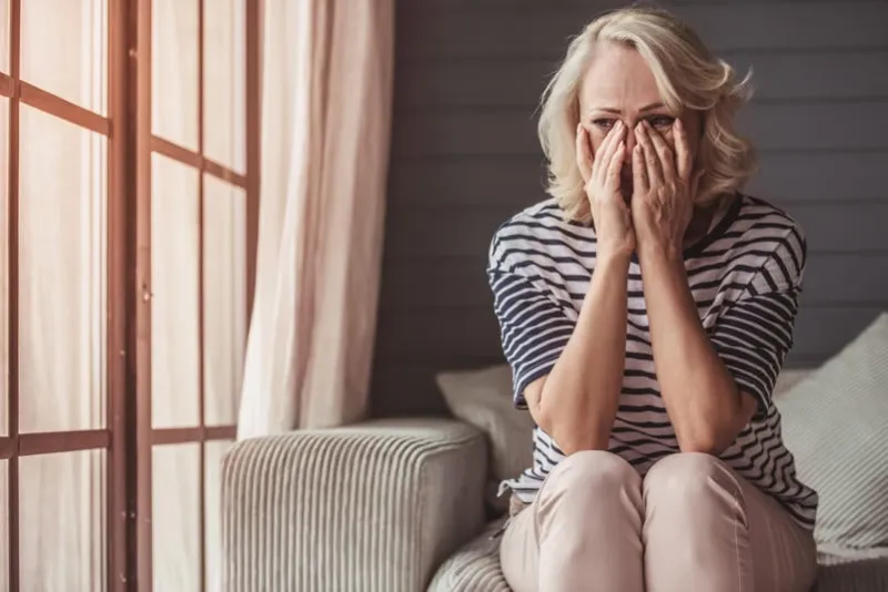 beautiful sad senior woman is crying and wiping her tears while sitting on couch at home