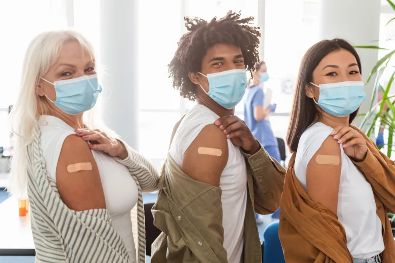 after vaccination concept portrait of happy vaccinated multicultural people in mask showing arm with sticking patch getting antiviral injection, sitting in line with rolled up sleeve healthcare