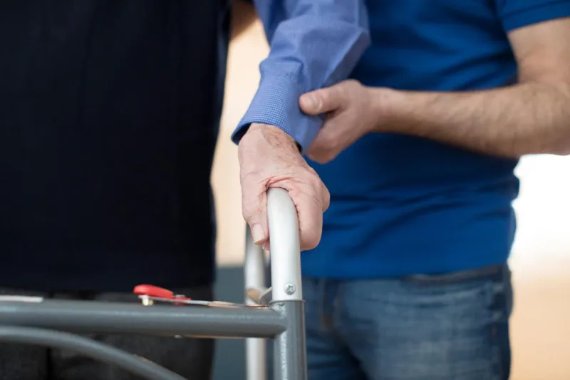 senior man's hands on walking frame with care worker in background