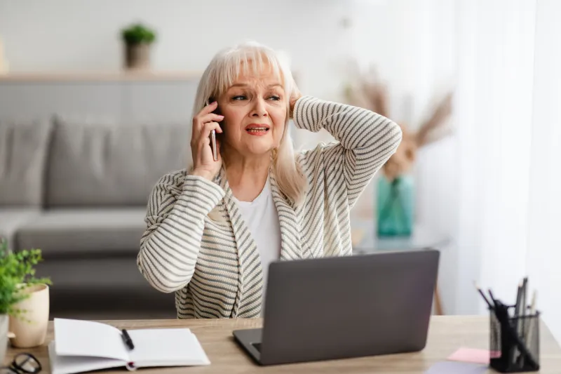 portrait of stressed senior lady making call using mobile phone near window in modern office fustrated mature woman having unpleasant conversation, showing negative emotion, grabbing head