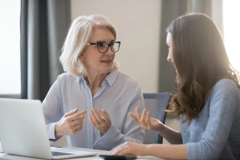 mature and young women colleagues sitting at desk talking about project startup ideas, sharing thoughts, solve currents issues, make research, discuss growth strategy, think how generate more revenue