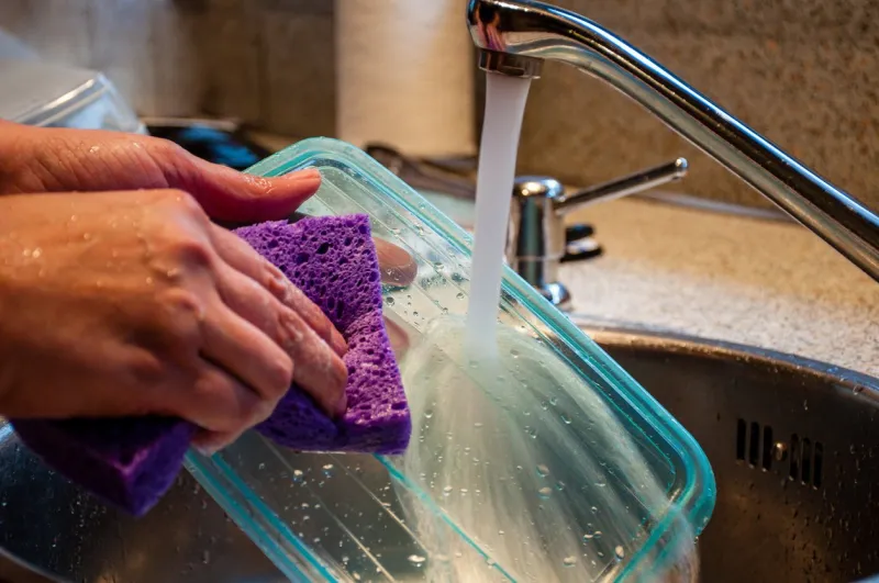 woman washing a plastic food storage container with a sponge in kitchen sink