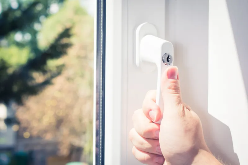 a male hand holding a lockable window handle of a closed white window with trees in background, prevent burglary concept