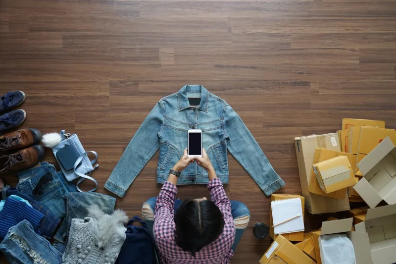top view of women take a photo jeans jecket with smart phone from home on wooden floor with postal parcel, selling online ideas concept